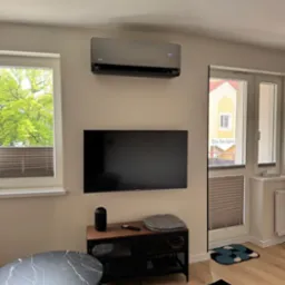 Szary klimatyzator VIMPA zamontowany nad telewizorem w jasnym pokoju dziennym z widokiem na zieleń za oknem. Minimalistyczny design wnętrza.