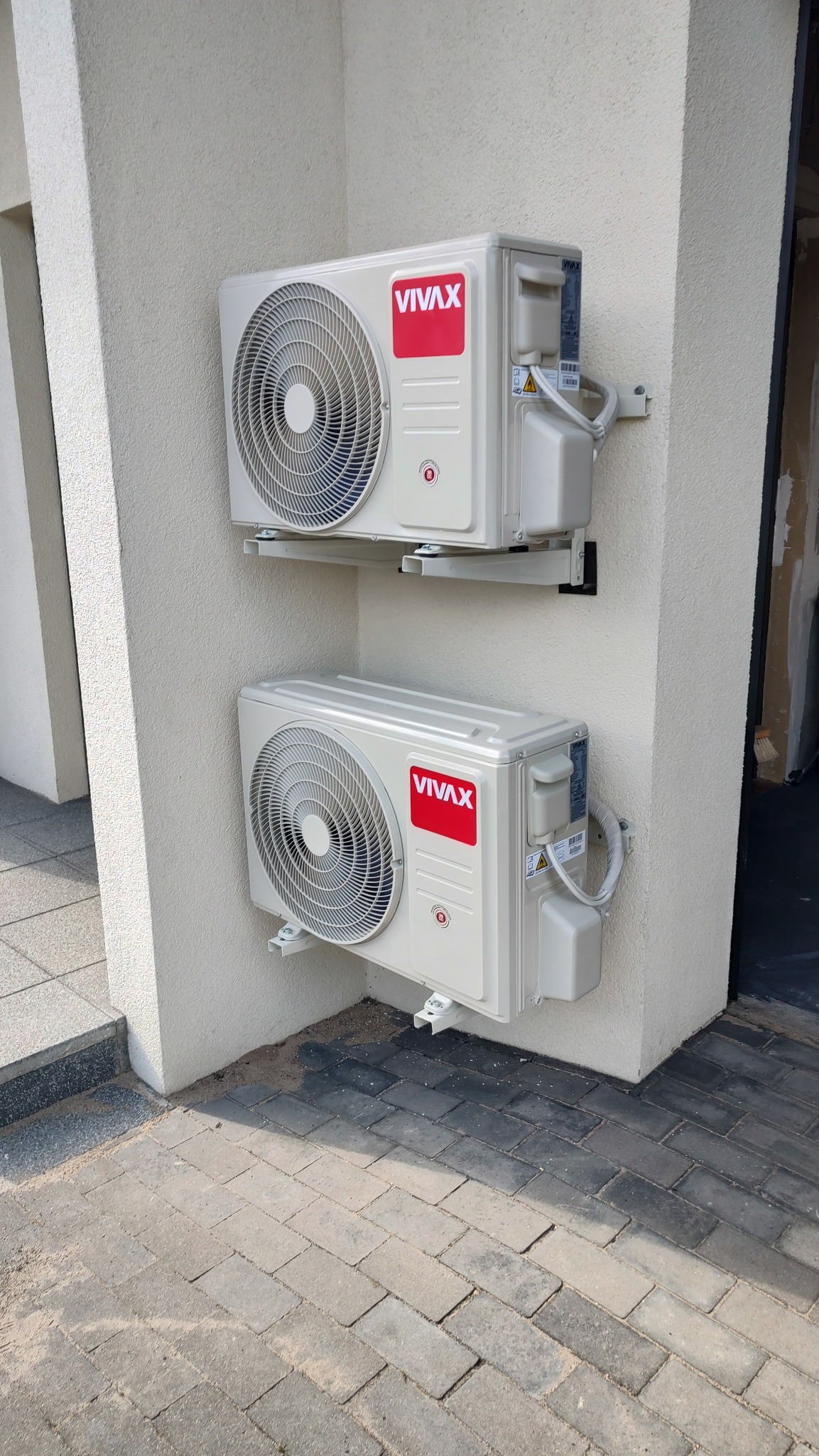 Dwa klimatyzatory VIVAX zamontowane jeden nad drugim na zewnętrznej ścianie budynku, widoczne przewody i uchwyty montażowe, kostka brukowa pod spodem.