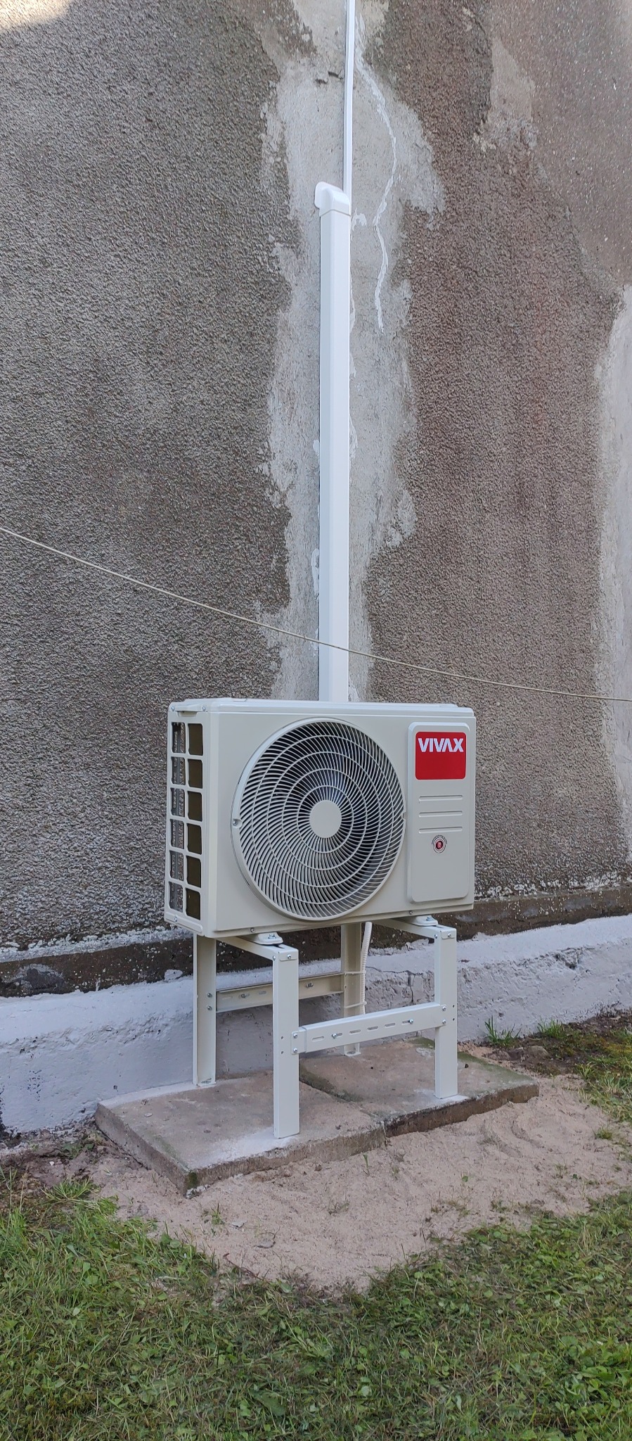Zewnętrzna jednostka klimatyzatora marki Vivax, zamontowana na metalowym stelażu przy szarej, chropowatej ścianie budynku, z widoczną rurą odprowadzającą skropliny.