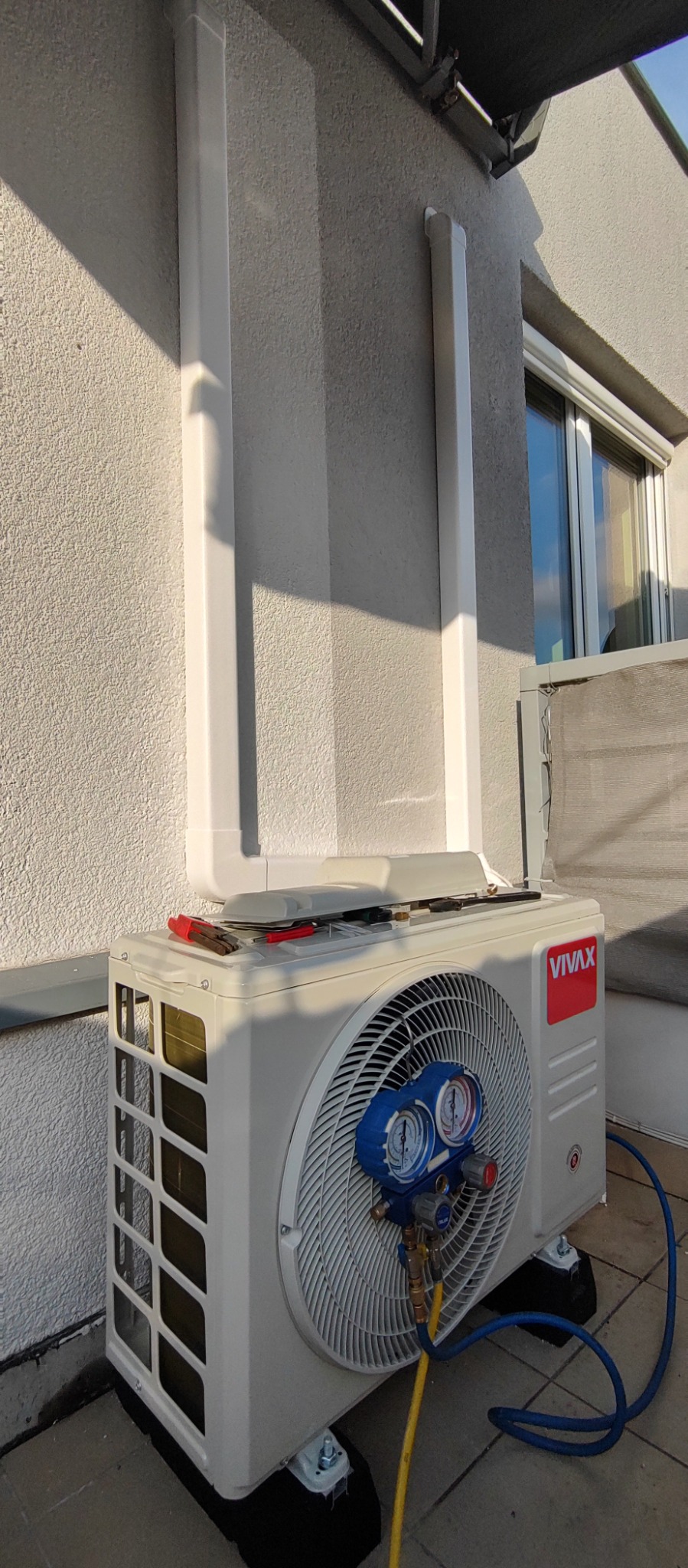 Zewnętrzna jednostka klimatyzacji marki Vivax z podłączonymi manometrami serwisowymi, umieszczona na balkonie, z widocznymi rurami odprowadzającymi skropliny i narzędziami na obudowie.