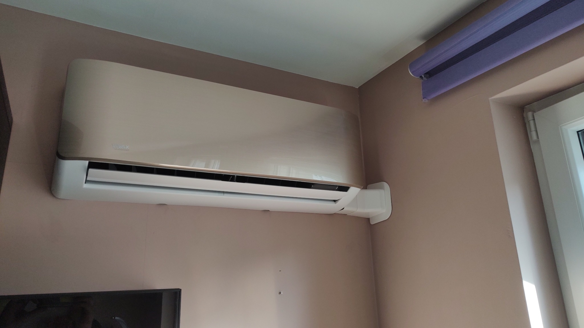 Beżowy klimatyzator VIVAX zamontowany na ścianie w pokoju z fioletową roletą okienną.