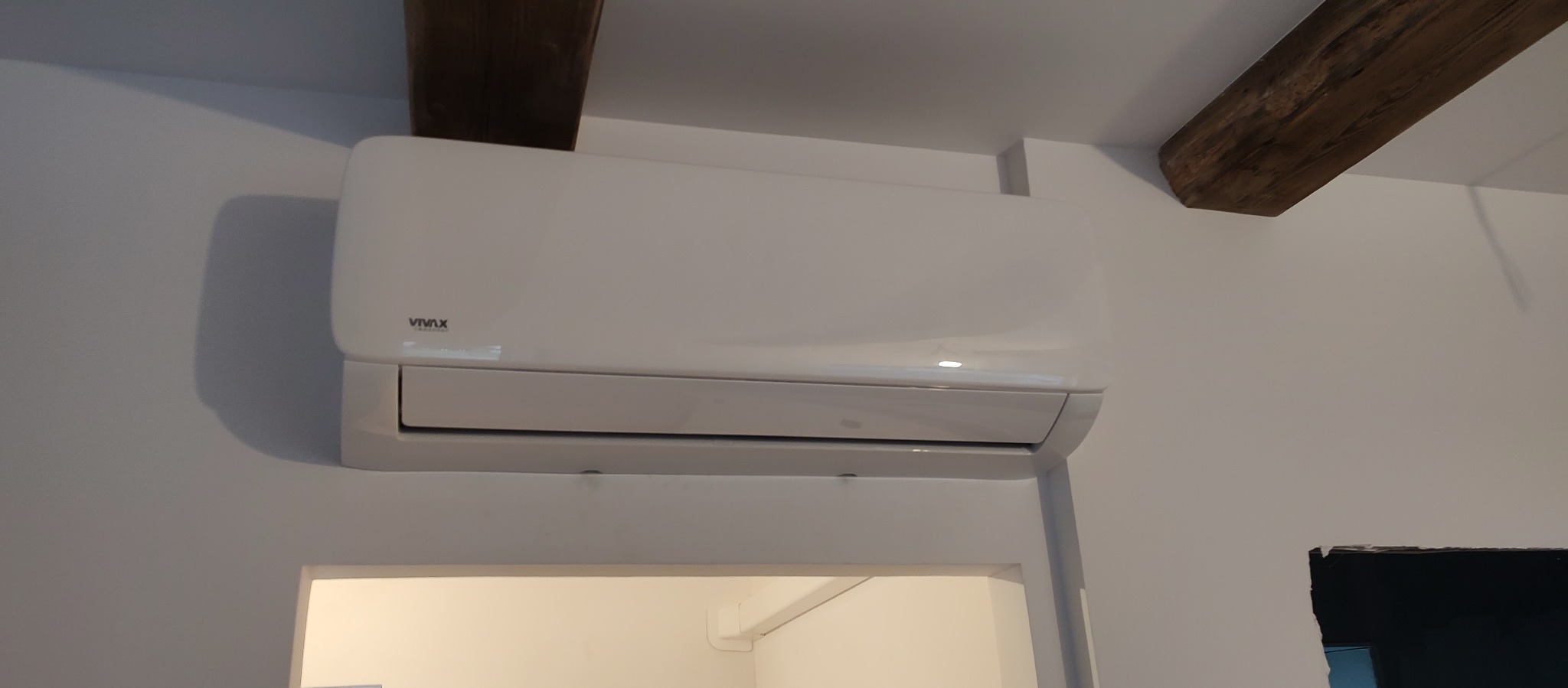 Biały klimatyzator marki Vivax zamontowany na ścianie w pomieszczeniu z drewnianymi belkami stropowymi.