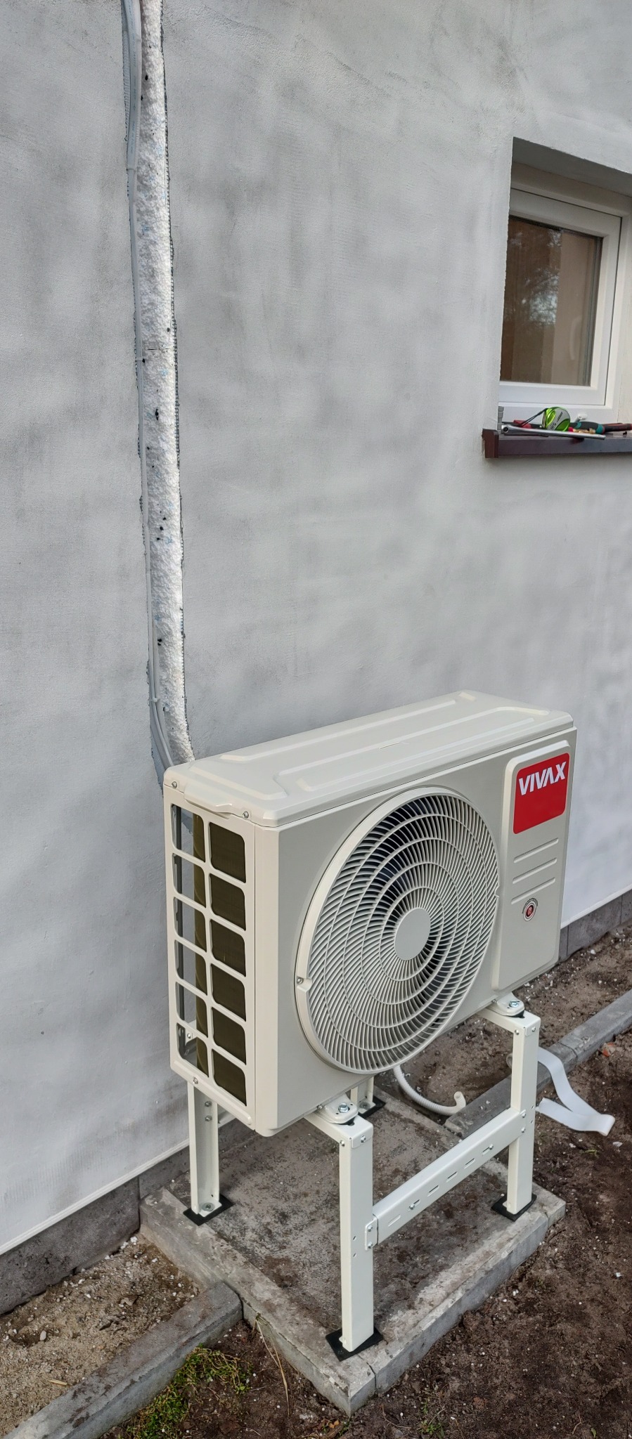 Zewnętrzna jednostka klimatyzacji VIVAX zamontowana na białym stelażu przy szarej ścianie budynku z widoczną instalacją w izolacji i narzędziami na parapecie okna.