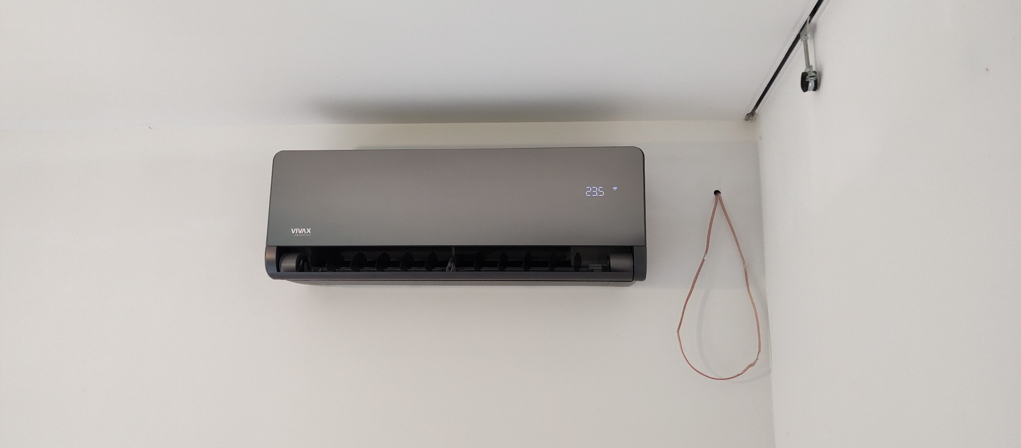 Szary klimatyzator VIVAX zamontowany na białej ścianie, widoczne wystające przewody elektryczne i wyświetlacz temperatury 23.5 stopnia.