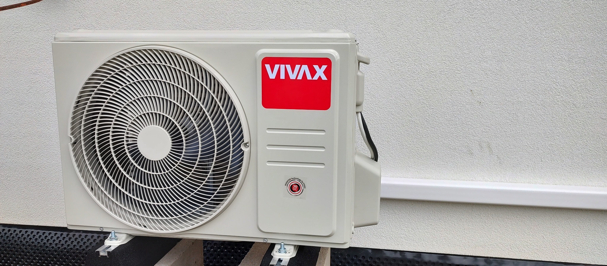 Zewnętrzna jednostka klimatyzacji VIVAX zamontowana na białej, teksturowanej ścianie budynku, z widocznymi przewodami w białej listwie przypodłogowej.