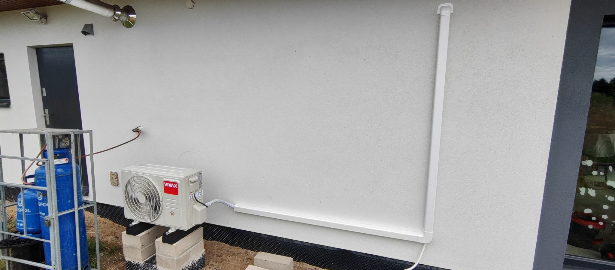 Zewnętrzna jednostka klimatyzatora Vivax zamontowana na betonowych blokach przy ścianie budynku, z widocznymi rurami odprowadzającymi oraz butlami z gazem w metalowej klatce obok.