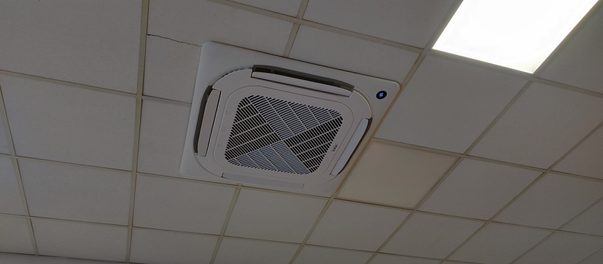 Kasetonowa klimatyzacja sufitowa VIVAX z widocznym wyświetlaczem temperatury, zamontowana w biurze. Widoczna kratka wentylacyjna i kasetonowy sufit.