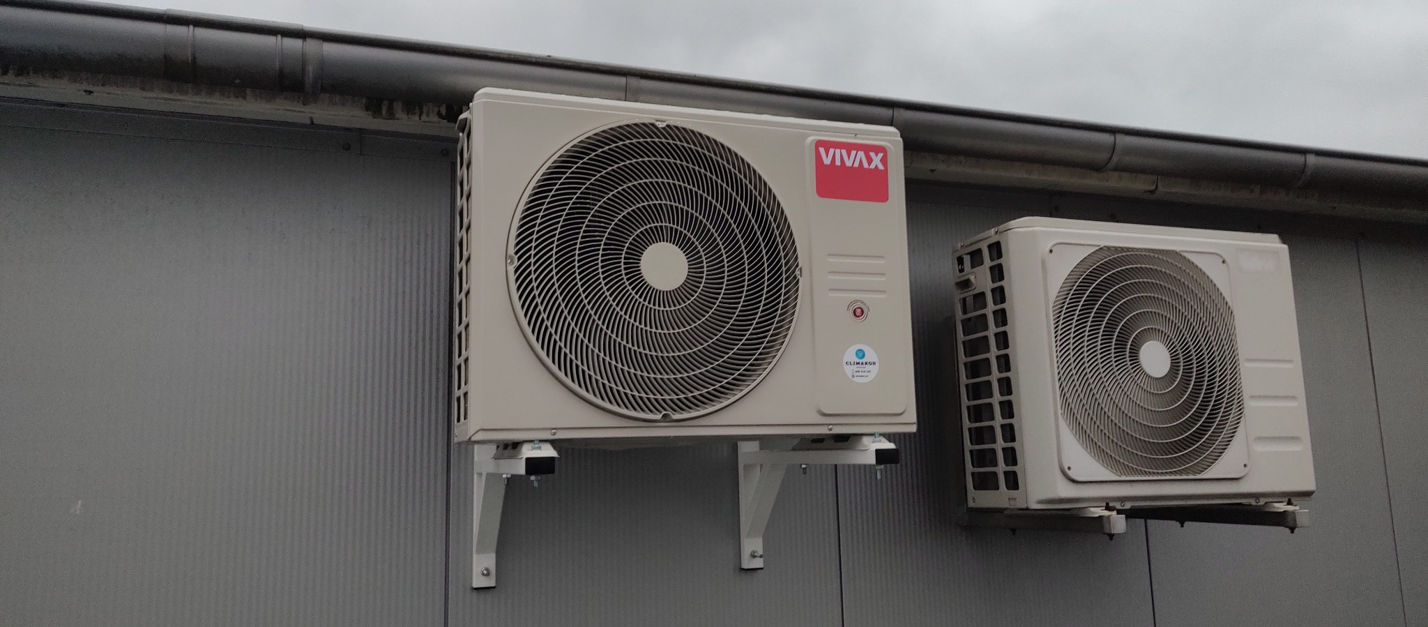 Dwa klimatyzatory VIVAX zamontowane na szarej, pionowo żłobkowanej ścianie budynku. Jeden z klimatyzatorów jest lekko przesunięty w lewo.