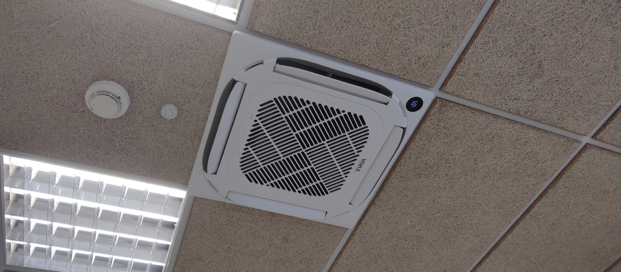 Kasetonowa klimatyzacja VIVAX zamontowana w suficie podwieszanym, obok czujnik dymu i oświetlenie. Temperatura na wyświetlaczu klimatyzacji to 16 stopni.