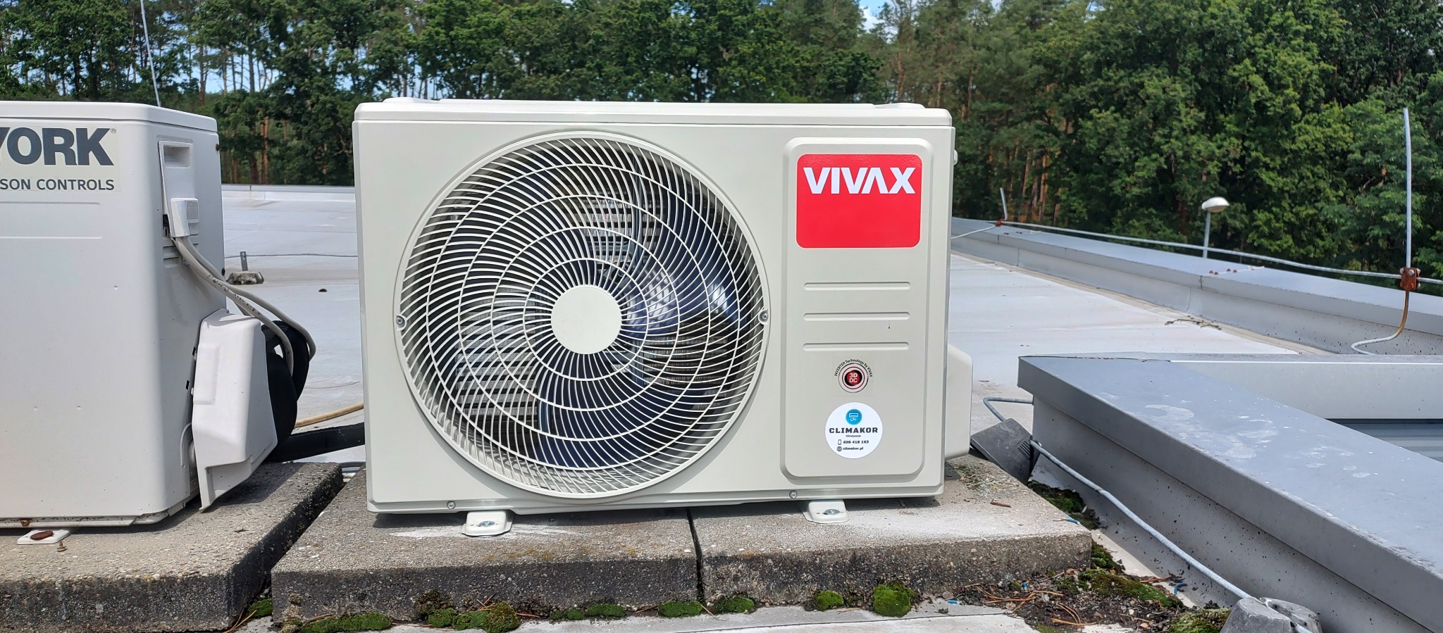 Zamontowana, beżowa jednostka zewnętrzna klimatyzacji Vivax na betonowej podstawie, z widocznym wentylatorem i logo firmy, na tle zielonych drzew.