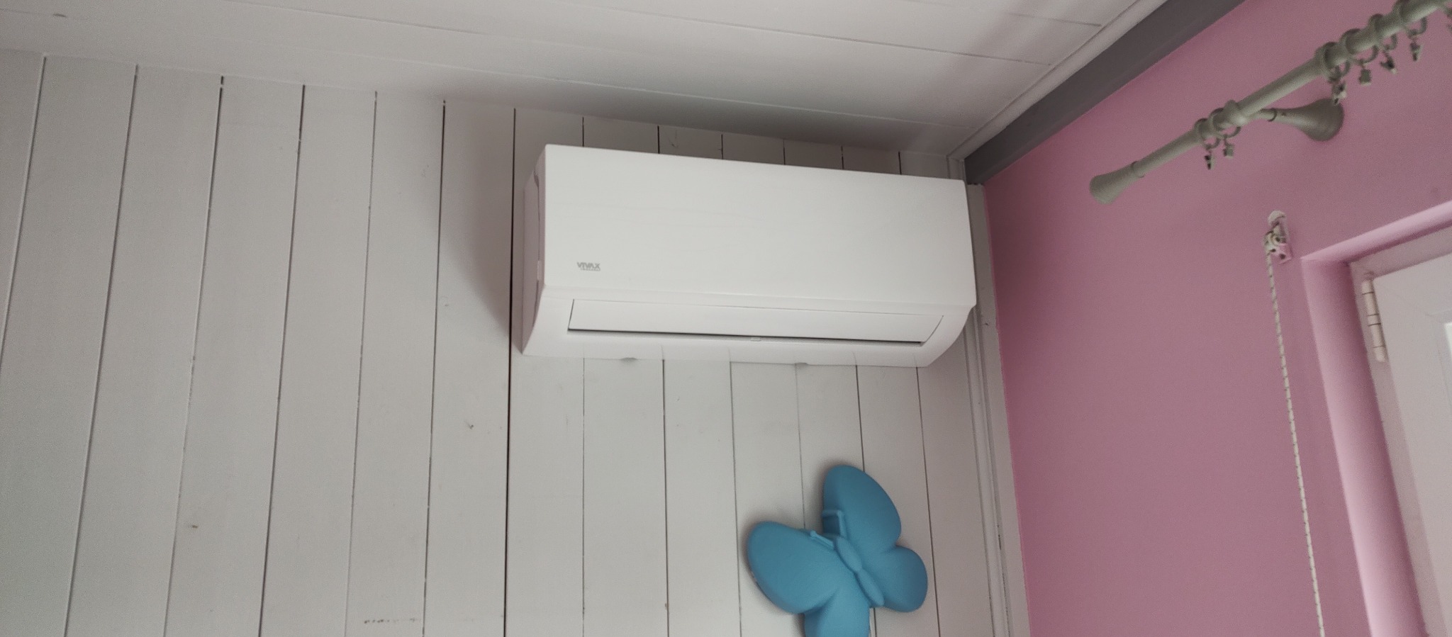 Biała klimatyzacja Vivax na ścianie z białych paneli, obok różowej ściany z karniszem i ozdobnym, błękitnym motylem poniżej.