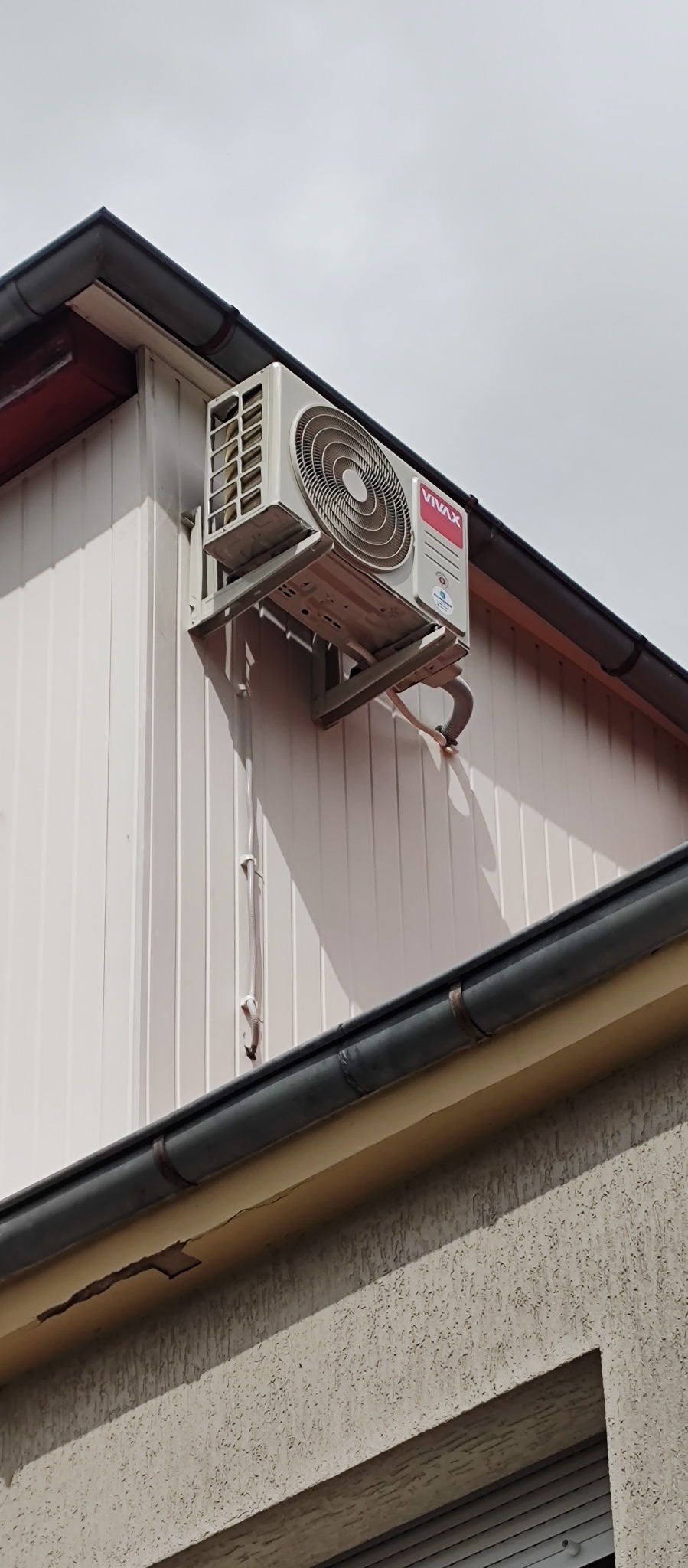 Klimatyzator VIVAX zamontowany na elewacji budynku z widocznymi przewodami instalacyjnymi. Ujęcie z dołu, pochmurne niebo w tle.