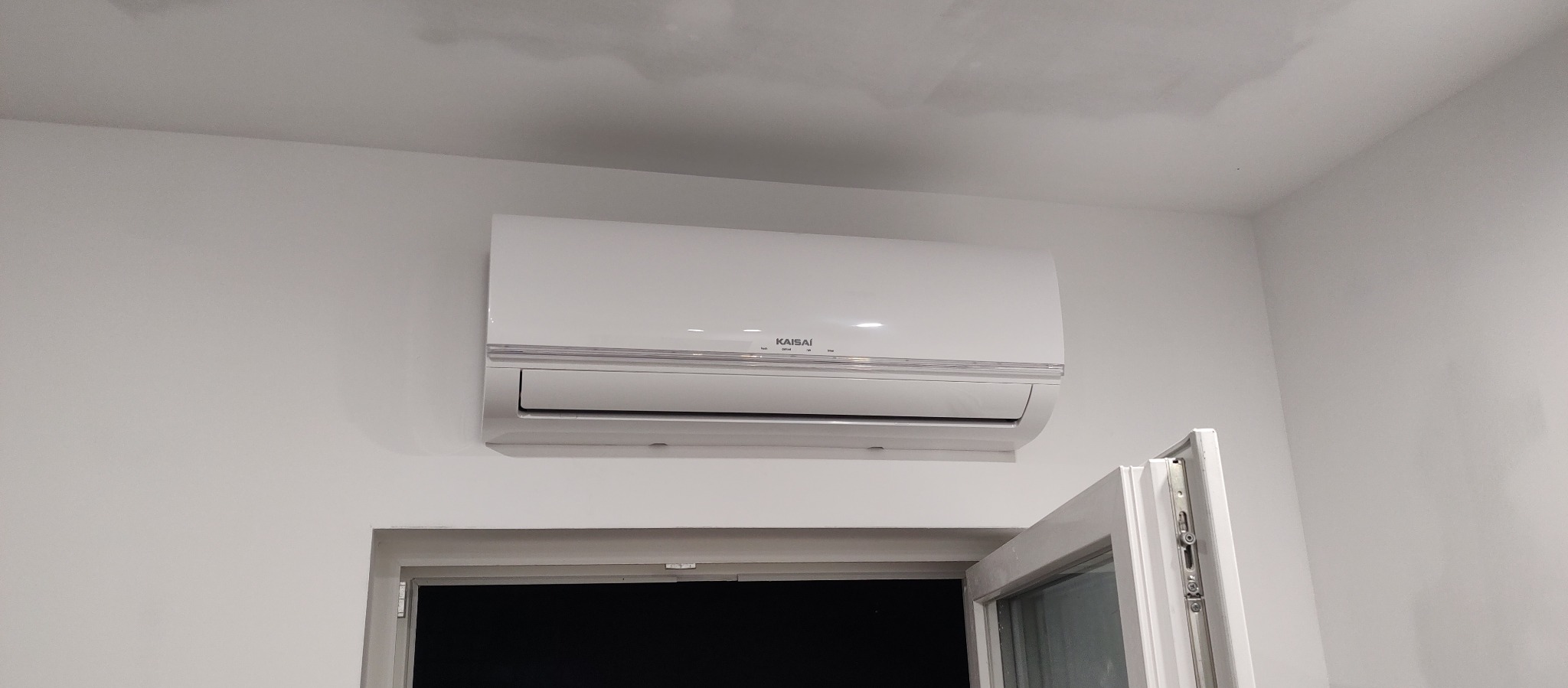 Zamontowana, biała klimatyzacja Kaisai na ścianie nad oknem. Widoczne detale urządzenia i otoczenie w jasnym wnętrzu. Minimalistyczny design.