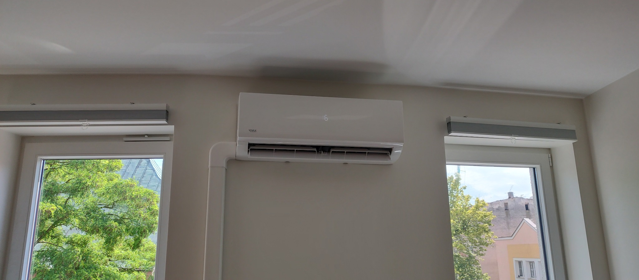 Biały klimatyzator Vivax zamontowany na ścianie w jasnym wnętrzu, widoczne okna z roletami i zielenią za nimi. Minimalistyczny design, estetyczny montaż.