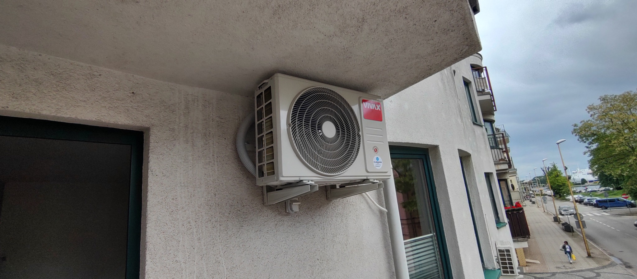 Biała jednostka zewnętrzna klimatyzacji VIVAX zamontowana na elewacji budynku mieszkalnego, widok z dołu, szary przewód odprowadzający skropliny.