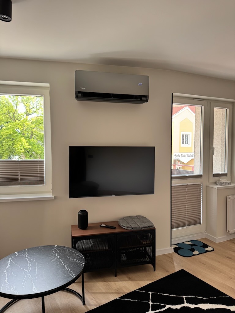 Szary klimatyzator VIMPA zamontowany nad telewizorem w jasnym pokoju dziennym z widokiem na zieleń za oknem. Minimalistyczny design wnętrza.