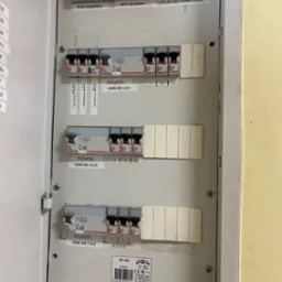 Otwarta skrzynka bezpiecznikowa z opisami obwodów w Świętochłowicach. Widoczne wyłączniki różnicowoprądowe i etykiety z numerami. Oświetlenie sufitowe.