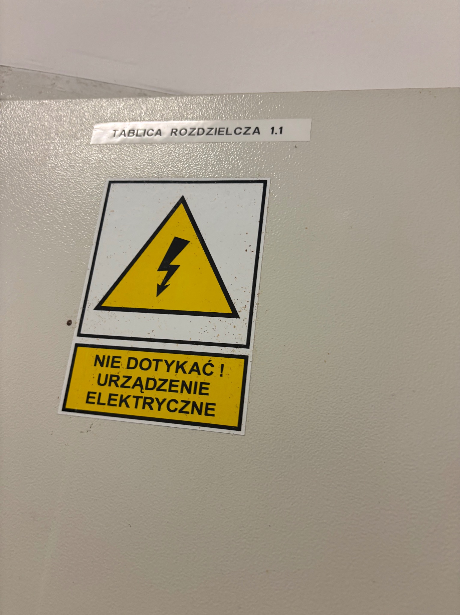 Tablica rozdzielcza z żółtą naklejką ostrzegawczą: 'Nie dotykać! Urządzenie elektryczne' i symbolem błyskawicy w trójkącie. Oznakowanie bezpieczeństwa.
