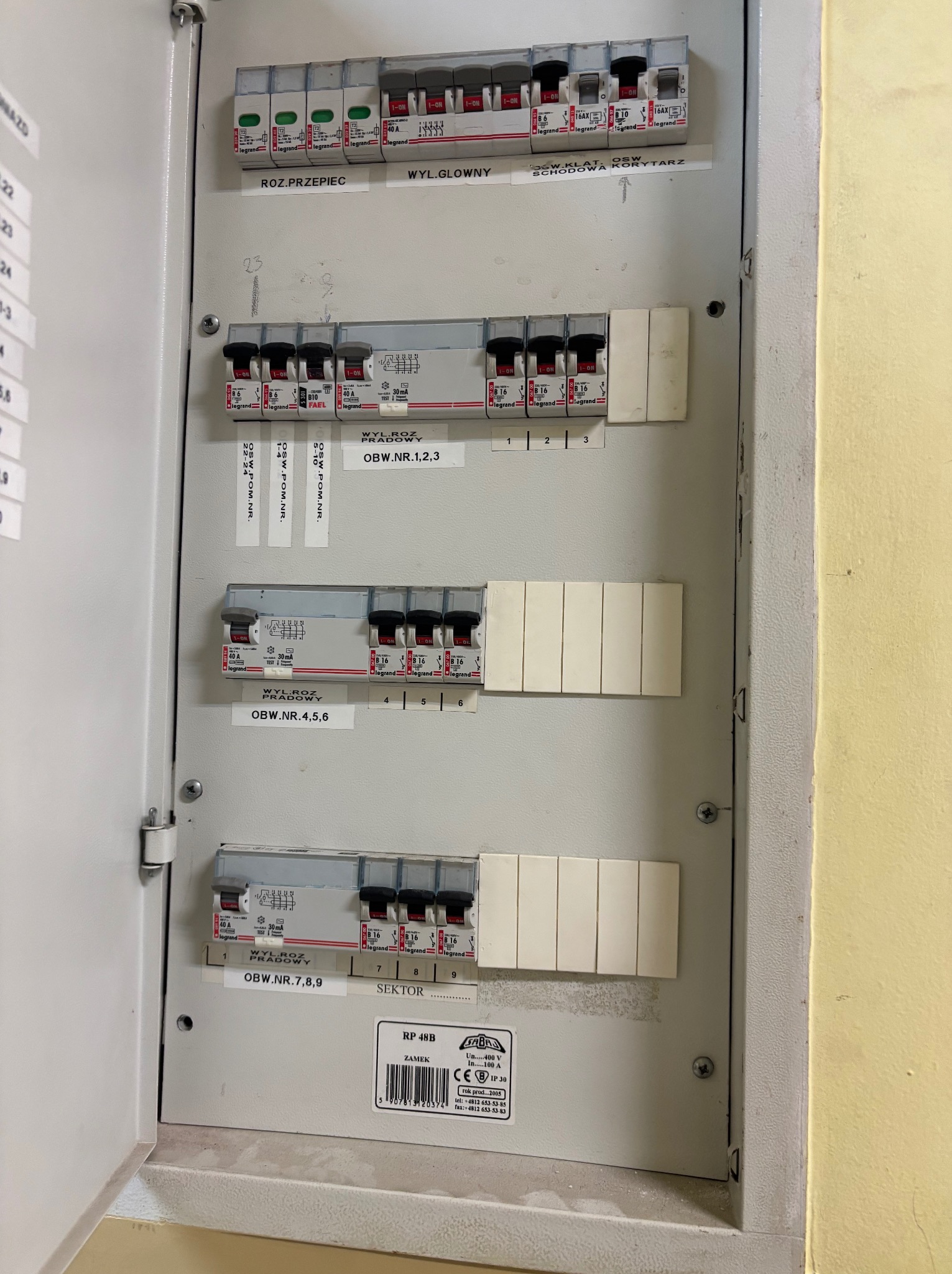 Otwarta skrzynka bezpiecznikowa z opisami obwodów w Świętochłowicach. Widoczne wyłączniki różnicowoprądowe i etykiety z numerami. Oświetlenie sufitowe.