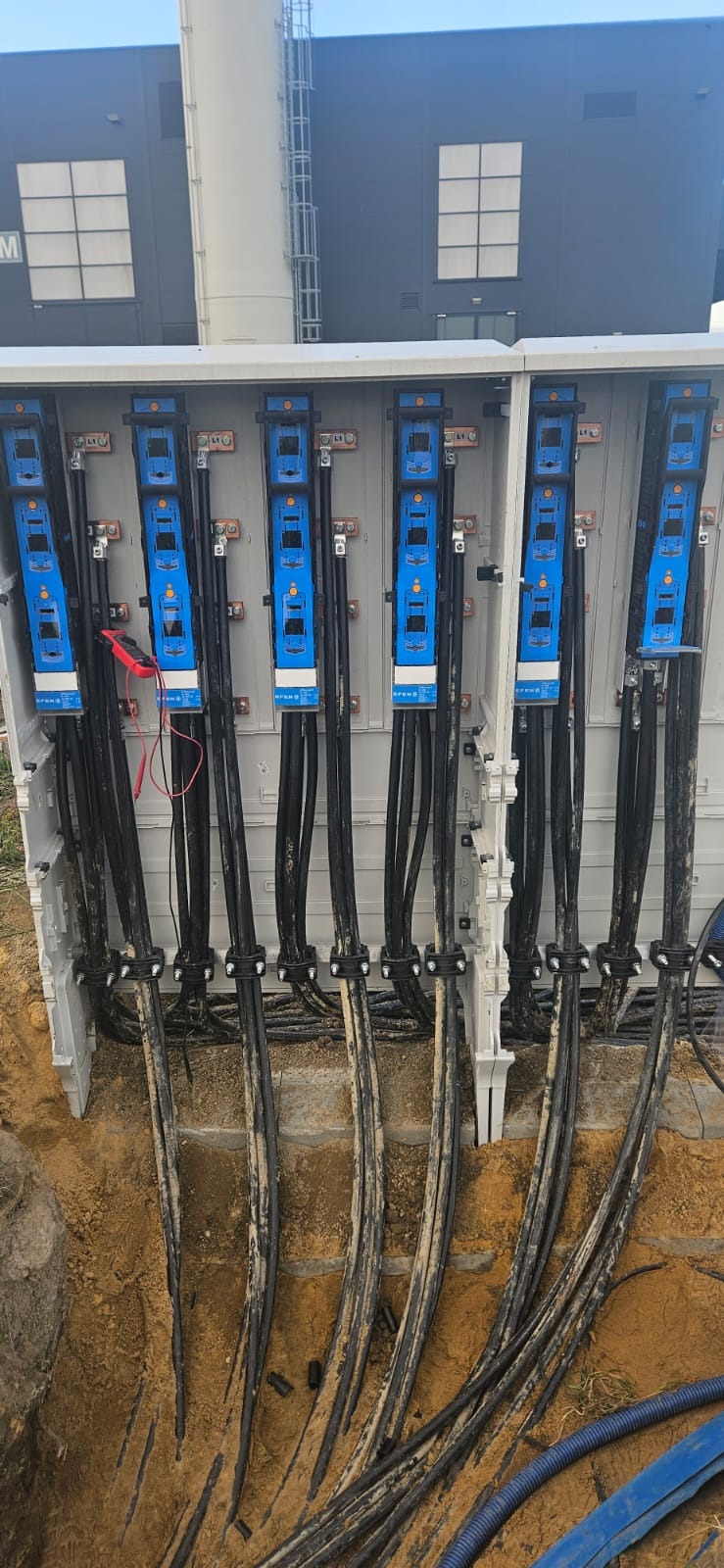 Otwarta skrzynka rozdzielcza z podłączonymi grubymi kablami w ziemi, widoczne mierniki elektryczne podłączone do jednego z modułów, budynek w tle.