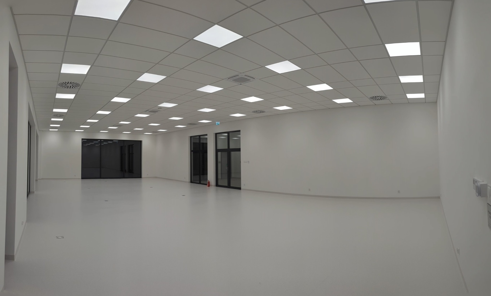 Wnętrze pustego, jasnego pomieszczenia z podwieszanym sufitem rastrowym i kwadratowymi oprawami oświetleniowymi, wentylacją mechaniczną i szklanymi drzwiami.