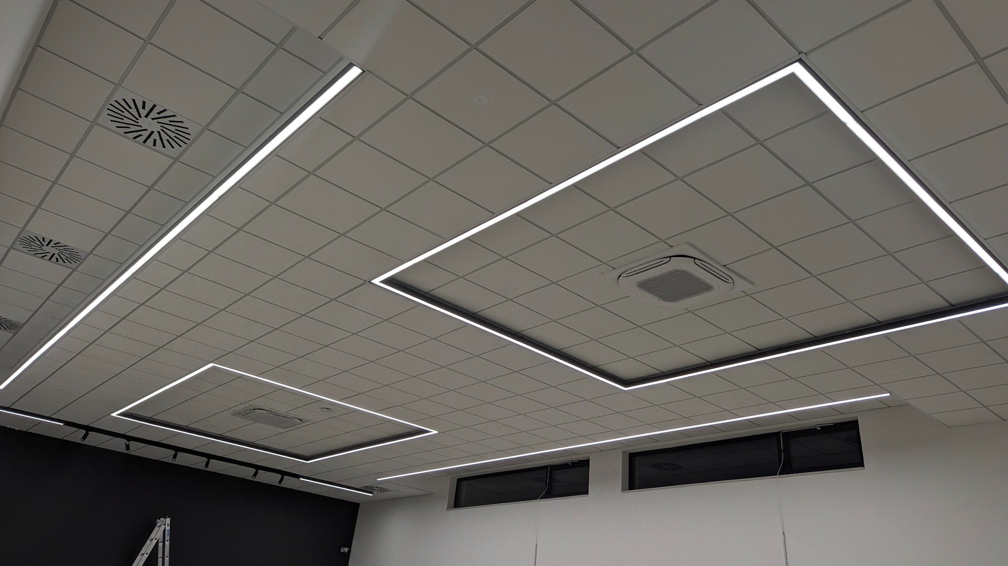 Sufit podwieszany z kwadratowymi panelami i liniowym oświetleniem LED tworzącym geometryczne wzory, widoczne kratki wentylacyjne i klimatyzatory kasetonowe.