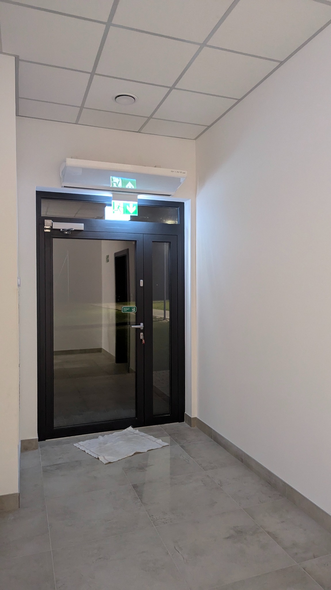 Dwuskrzydłowe, czarne drzwi ewakuacyjne z zielonym podświetlanym znakiem wyjścia awaryjnego nad nimi, widok z korytarza wyłożonego szarą terakotą.