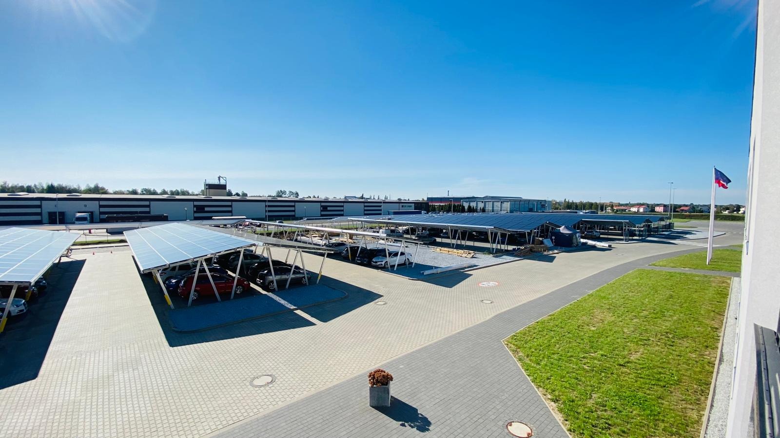 Zewnętrzny parking z zadaszeniem z paneli słonecznych, widok z góry w słoneczny dzień, budynek przemysłowy w tle.