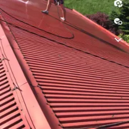 Pracownik na czerwonym, blaszanym dachu czyści go myjką ciśnieniową; w tle zieleń drzew.