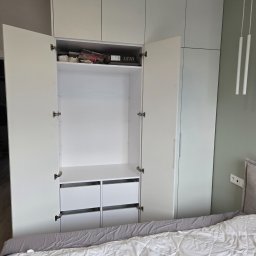DashPol - Nowoczesna, biała szafa wnękowa sięgająca sufitu, z otwartymi drzwiami ukazującymi półki i dwie szuflady, w sypialni z zieloną ścianą i minimalistycznymi lampami wiszącymi.