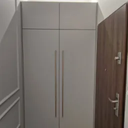 Szara szafa wnękowa z długimi, pionowymi uchwytami, przylegająca do ściany z dekoracyjną listwą i sąsiadująca z ciemnymi drzwiami wejściowymi.