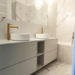 Elegancka łazienka z dwoma białymi umywalkami nablatowymi i złotymi bateriami, szare szafki pod umywalkami z otwartymi półkami, marmurowe płytki na ścianach i podłodze, oświetlenie punktowe...