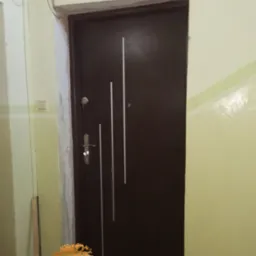 Ciemnobrązowe drzwi z trzema białymi, pionowymi paskami, zamontowane w odnowionym, ale niedokończonym wnętrzu, widoczne ślady po malowaniu na ścianach.