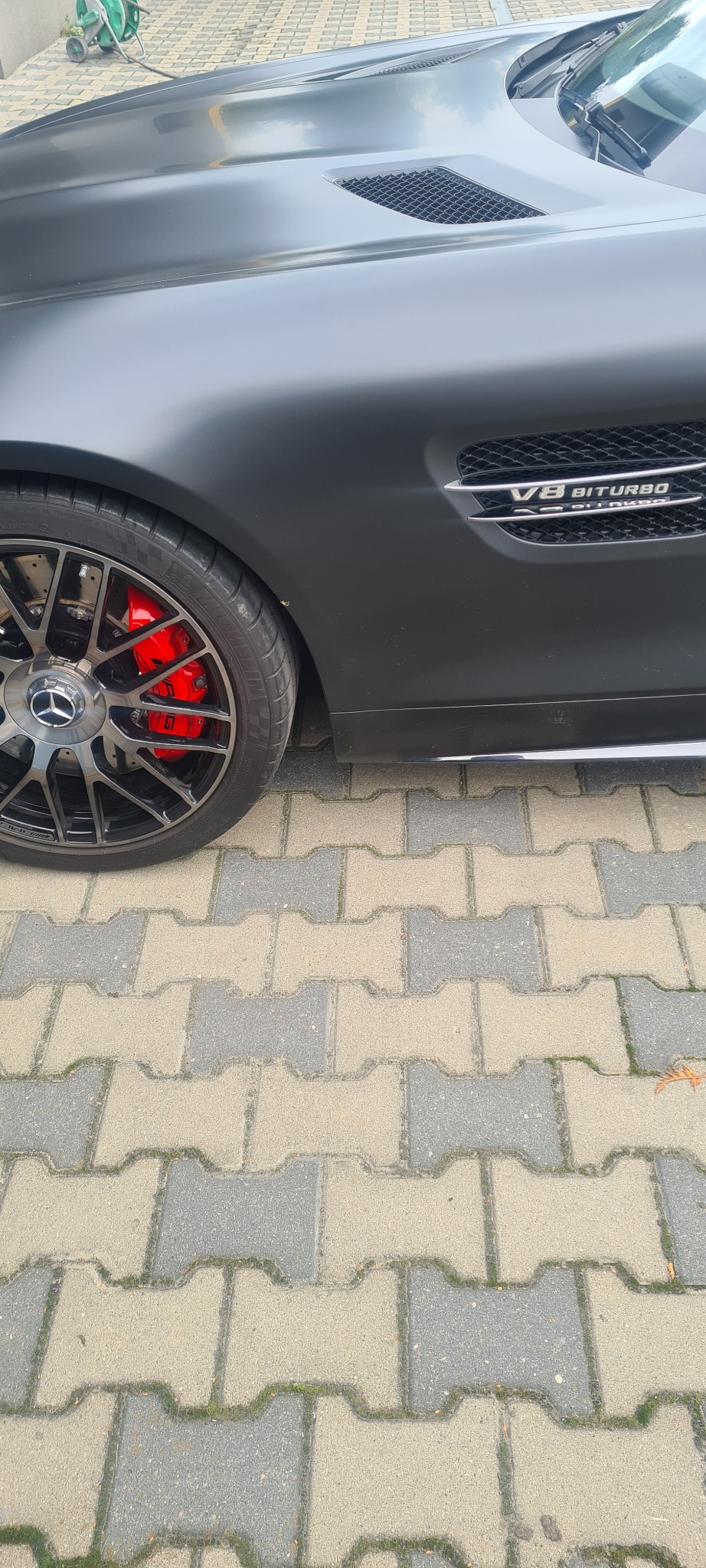 Matowy, szary samochód sportowy Mercedes-AMG z widocznymi czerwonymi zaciskami hamulcowymi i oznaczeniem V8 BITURBO na błotniku, zaparkowany na szarej kostce brukowej.