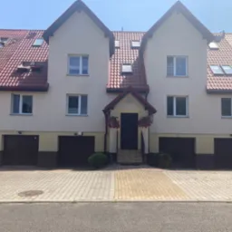 Trzy segmenty bliźniacze w Jawiszowicach z czerwonym dachem i jasną elewacją, widok z poziomu ulicy na front budynku z bramami garażowymi i wejściem ozdobionym wiszącymi kwiatami.