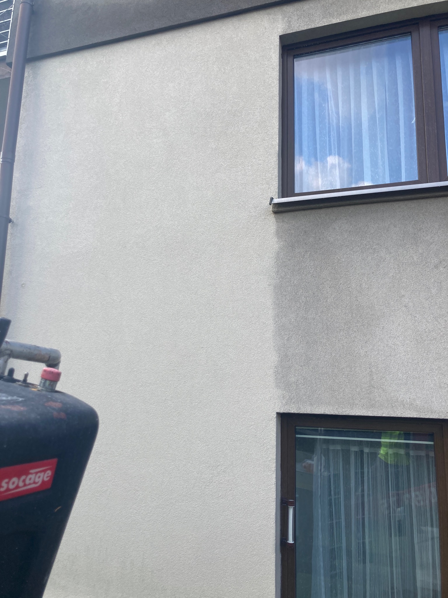 Fragment elewacji budynku z dwoma oknami i urządzeniem czyszczącym z widocznym logo 'Socage', sugerujący proces czyszczenia.