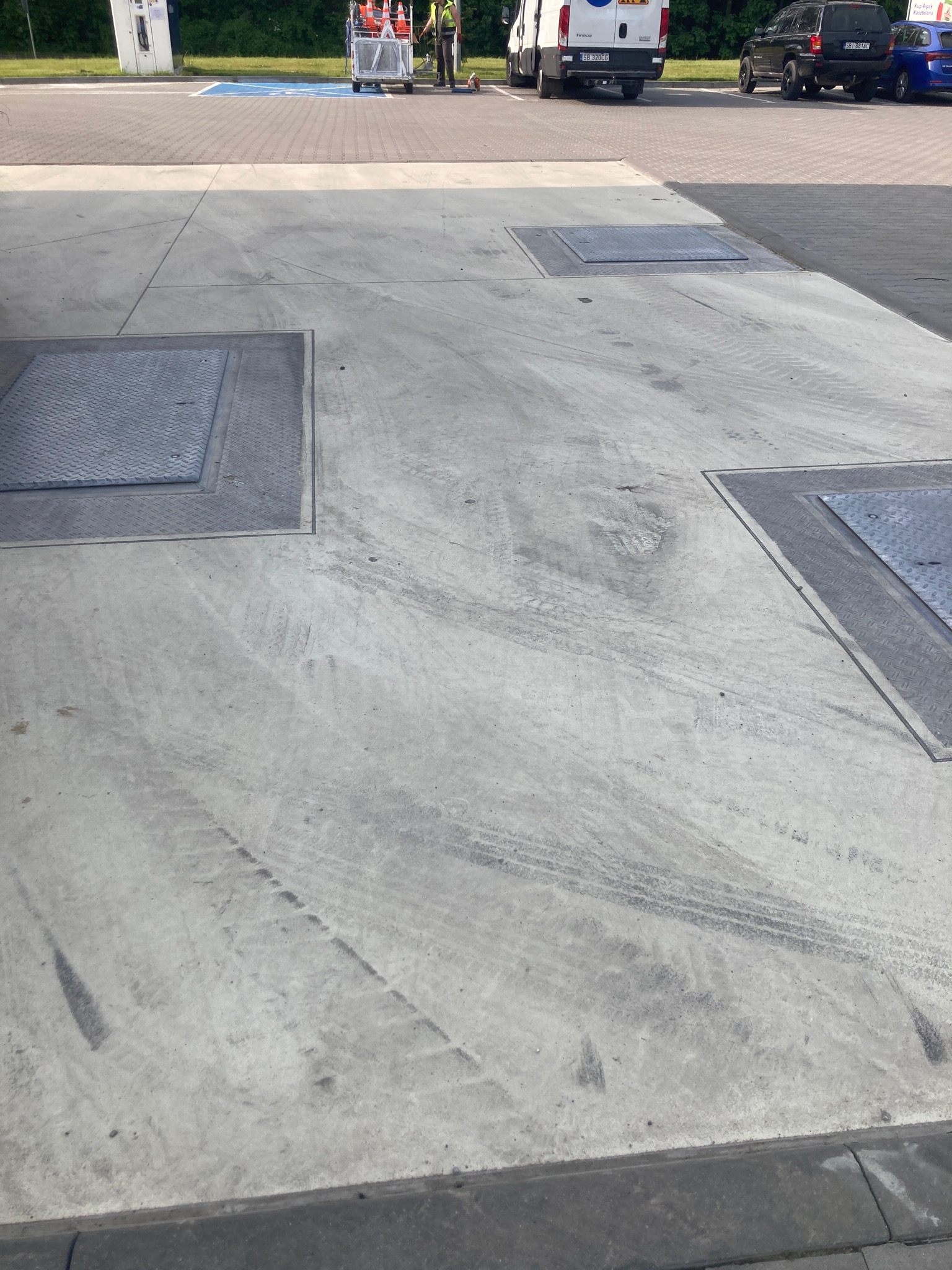 Betonowa nawierzchnia z metalowymi włazami i śladami opon, widoczni pracownicy z wózkiem i pachołkami w tle.