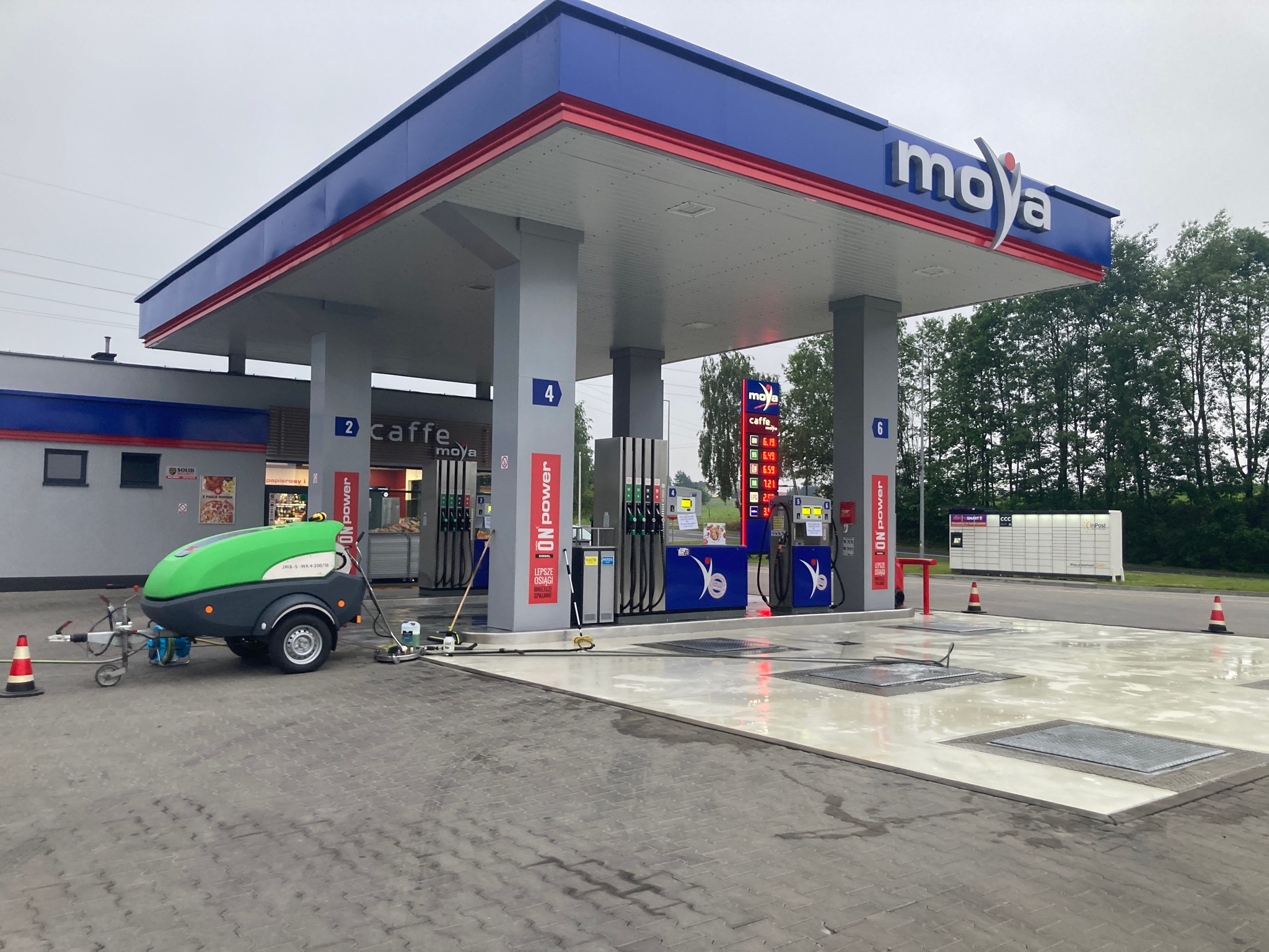 Stacja paliw Moya: świeżo umyta kostka brukowa na podjeździe, widoczny zielony wózek z urządzeniem do czyszczenia pod ciśnieniem i mokre plamy po myciu.