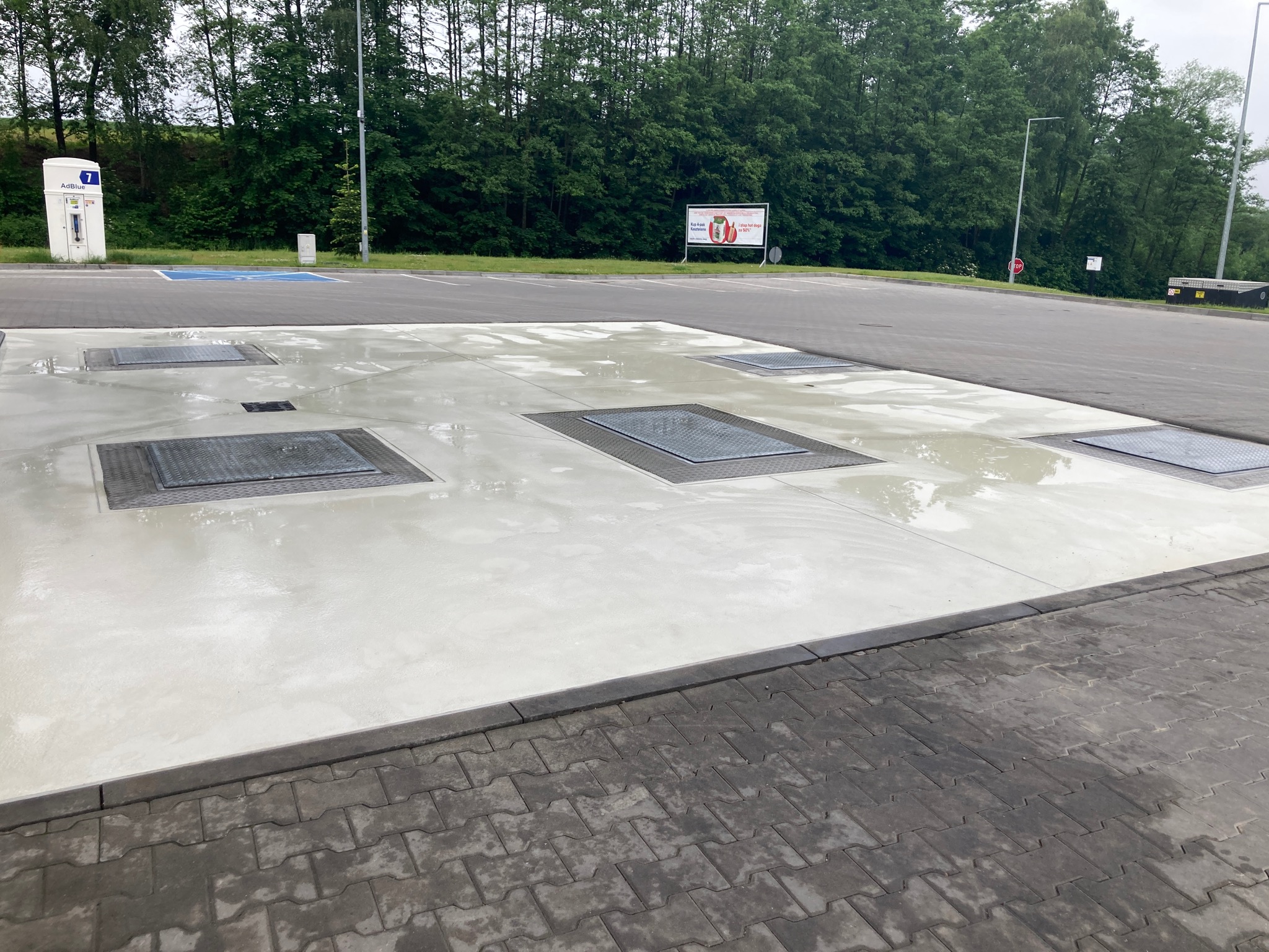 Świeżo umyta, betonowa powierzchnia z metalowymi włazami kanalizacyjnymi na stacji benzynowej, obok kostki brukowej i dystrybutora AdBlue.