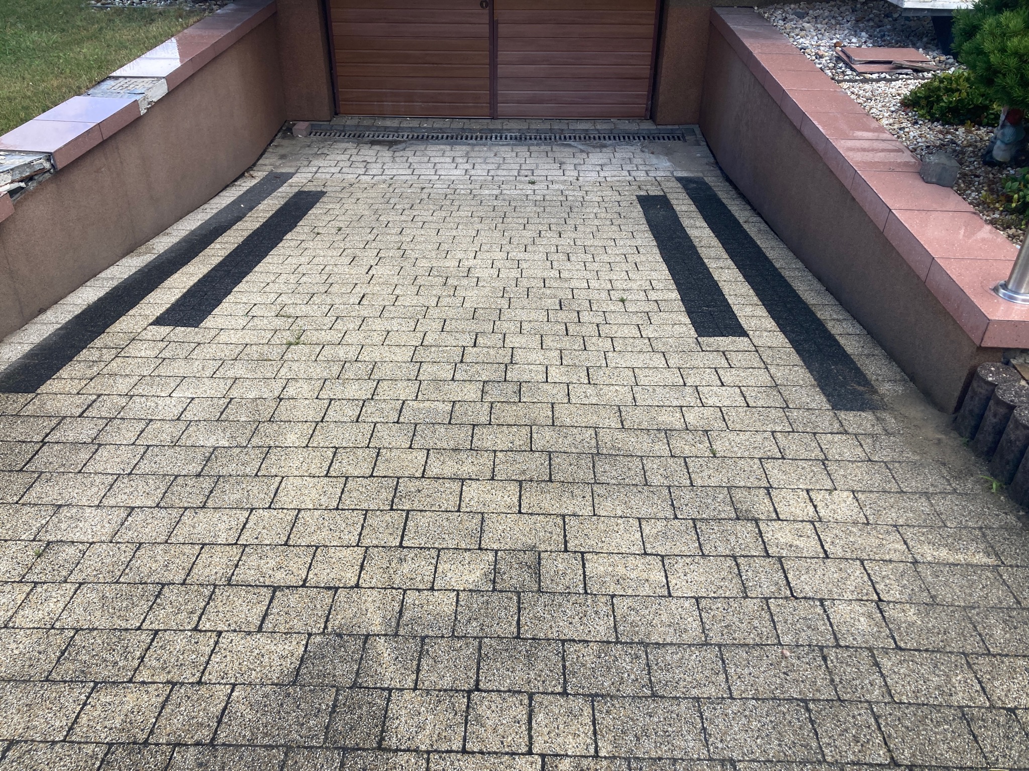 Szara kostka brukowa na podjeździe do garażu z brązowymi drzwiami, widoczne ciemniejsze pasy na kostce i fragmenty zieleni przy krawędziach.