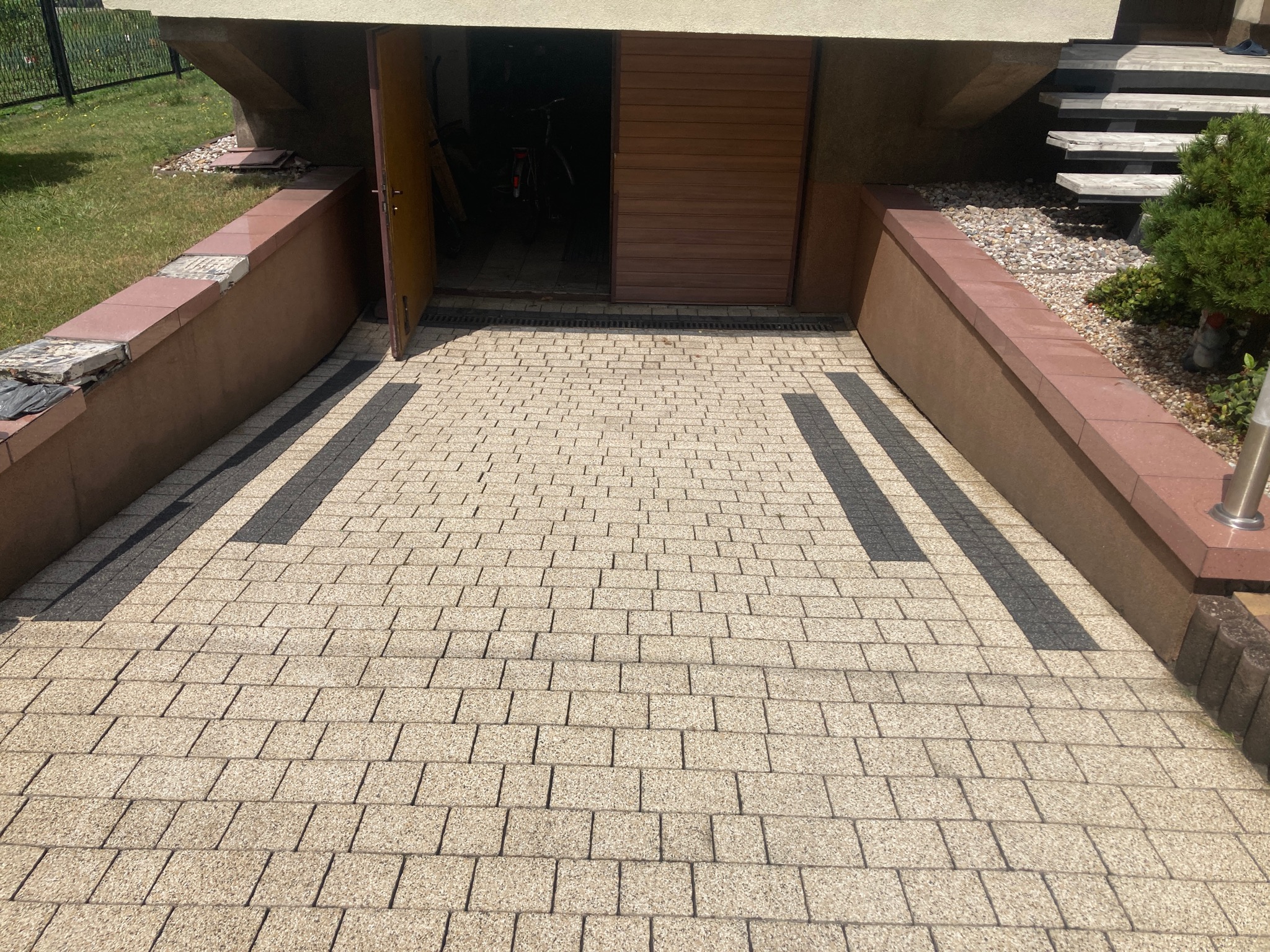 Jasna kostka brukowa przed garażem, z widocznymi ciemnymi pasami, otwartymi drzwiami garażowymi i fragmentami roślinności po bokach.