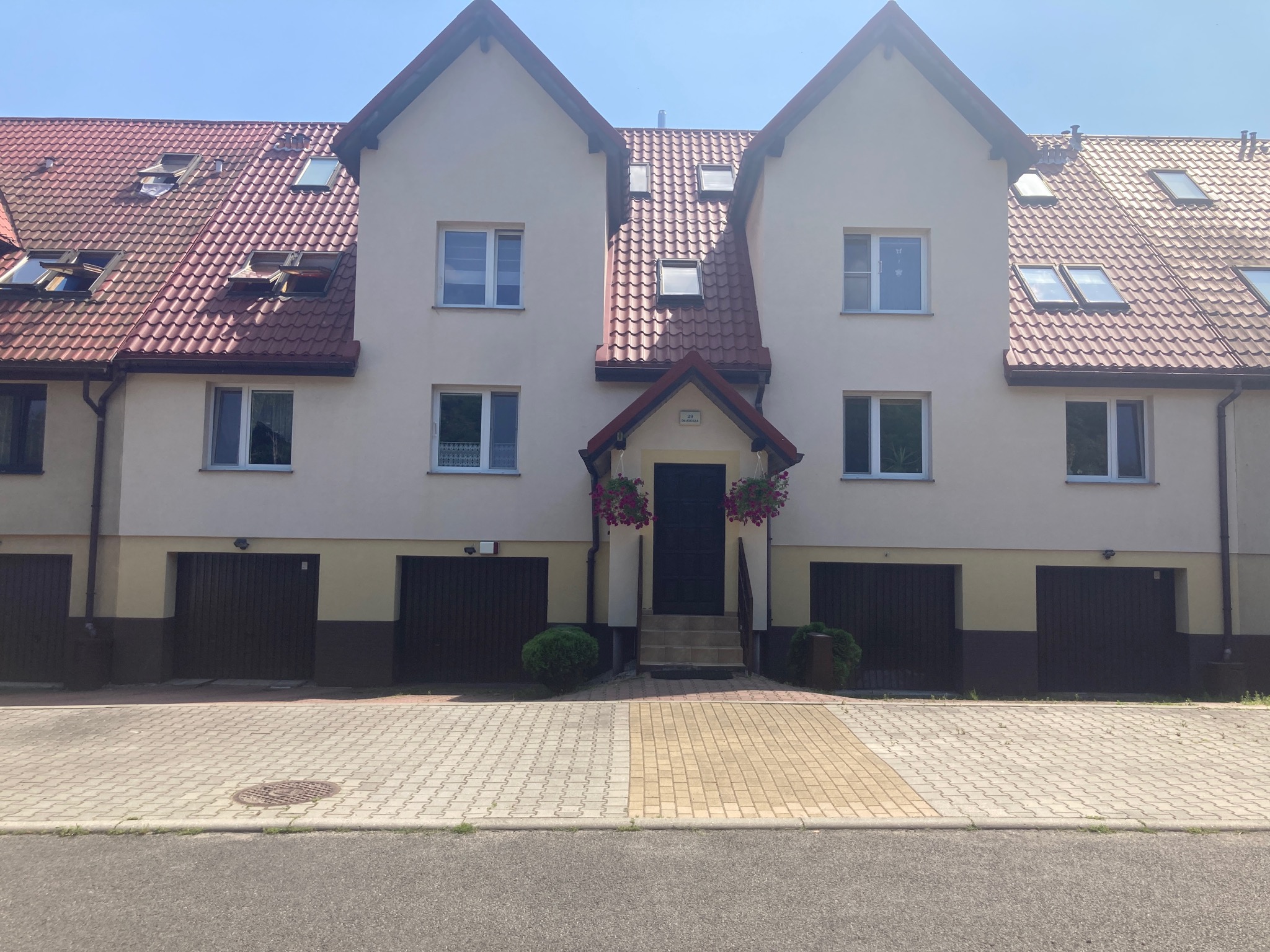 Trzy segmenty bliźniacze w Jawiszowicach z czerwonym dachem i jasną elewacją, widok z poziomu ulicy na front budynku z bramami garażowymi i wejściem ozdobionym wiszącymi kwiatami.