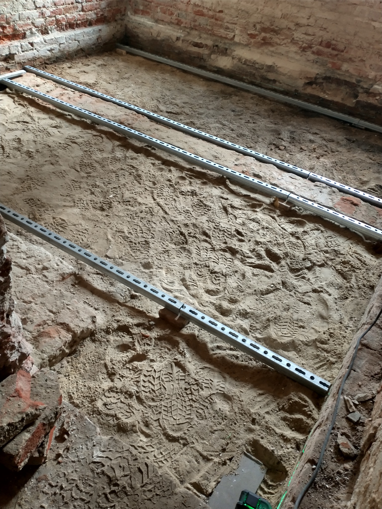 Przygotowanie podłoża pod posadzkę: stalowe profile ułożone na piasku z licznymi śladami butów, widoczne fragmenty ceglanej ściany i poziomica laserowa.