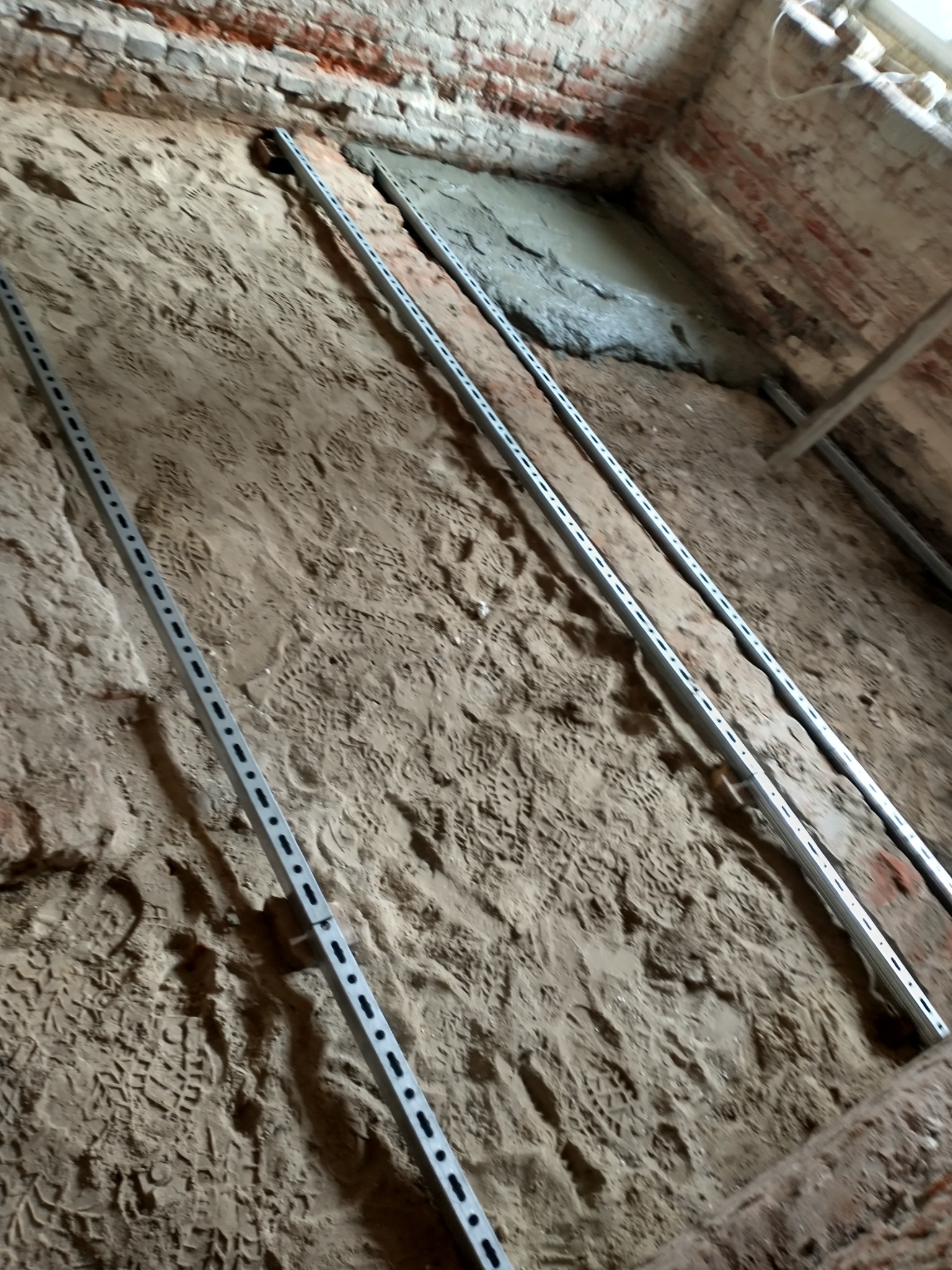 Przygotowanie podłoża pod wylewkę: widoczne metalowe profile poziomujące na piasku z licznymi śladami butów, fragment wylanej zaprawy i surowe ceglane ściany.