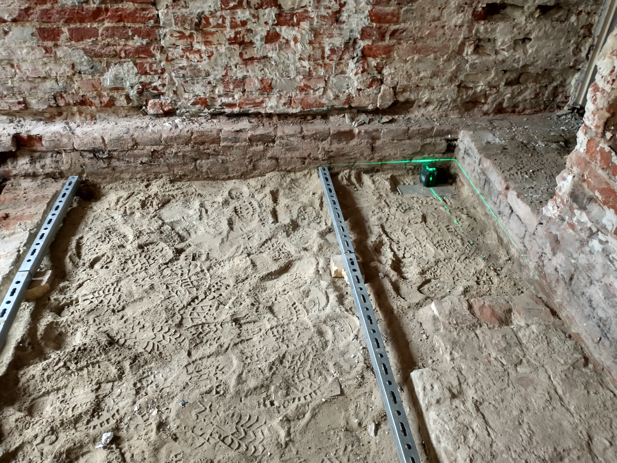 Przygotowanie podłoża pod posadzkę w pomieszczeniu z ceglanymi ścianami, widoczne metalowe profile, piasek z odciskami butów i laser krzyżowy wyznaczający poziom.