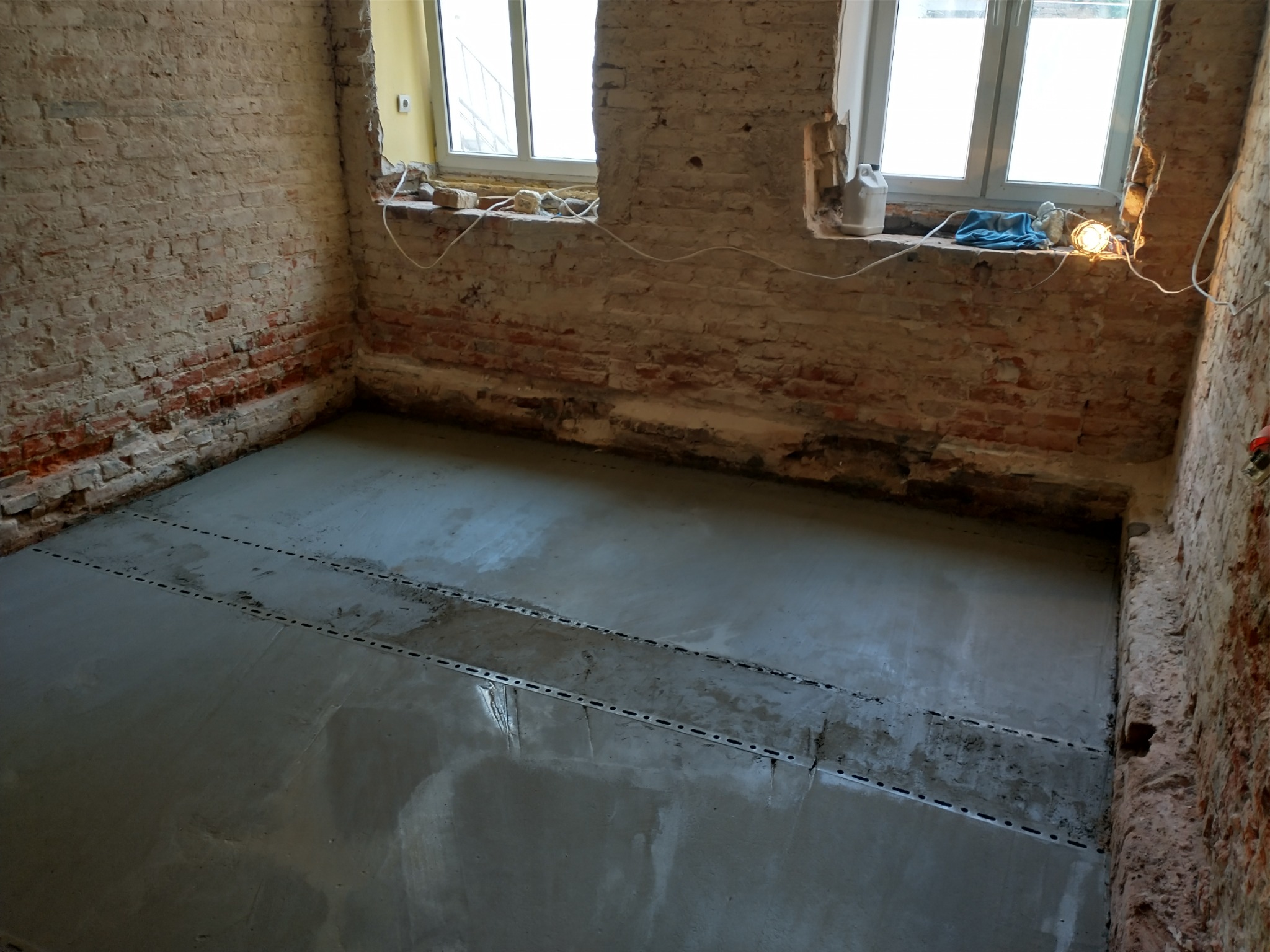 Świeżo wylana, mokra wylewka podłogowa ze stalowymi listwami dylatacyjnymi w surowym pomieszczeniu z odsłoniętą cegłą i oknami.