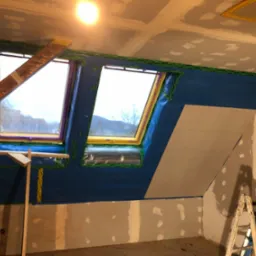 Poddasze w trakcie wykańczania: zamontowane okna dachowe zabezpieczone folią, częściowo położona płyta karton gips na skosie, ściany przygotowane do szpachlowania, widoczna drabina.