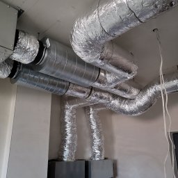 J.W Instal Tomasz Bohm - System kanałów wentylacyjnych z izolacją aluminiową, zamontowany pod sufitem w surowym stanie, widoczne przewody elektryczne i elementy konstrukcyjne budynku.