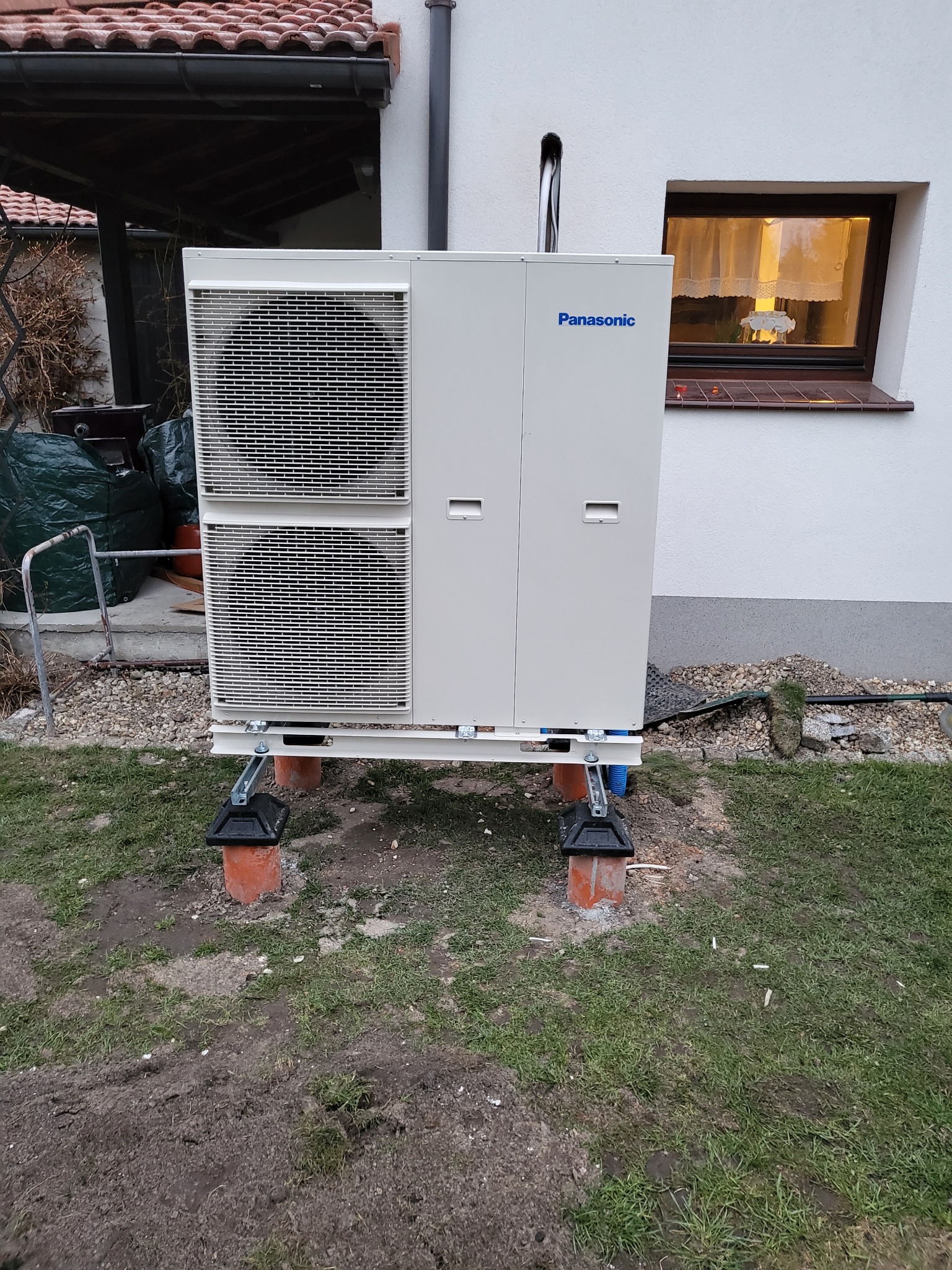 Zewnętrzna jednostka pompy ciepła Panasonic, zamontowana na podwyższeniu z cegieł i plastikowych elementów, z widocznym przewodem wychodzącym z góry, na tle ściany domu z oknem.