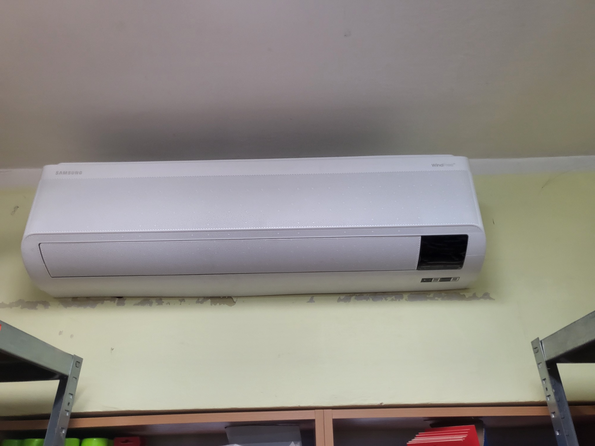 Biała jednostka wewnętrzna klimatyzatora Samsung WindFree zamontowana na ścianie nad regałem magazynowym, widoczne ślady po wcześniejszym malowaniu.